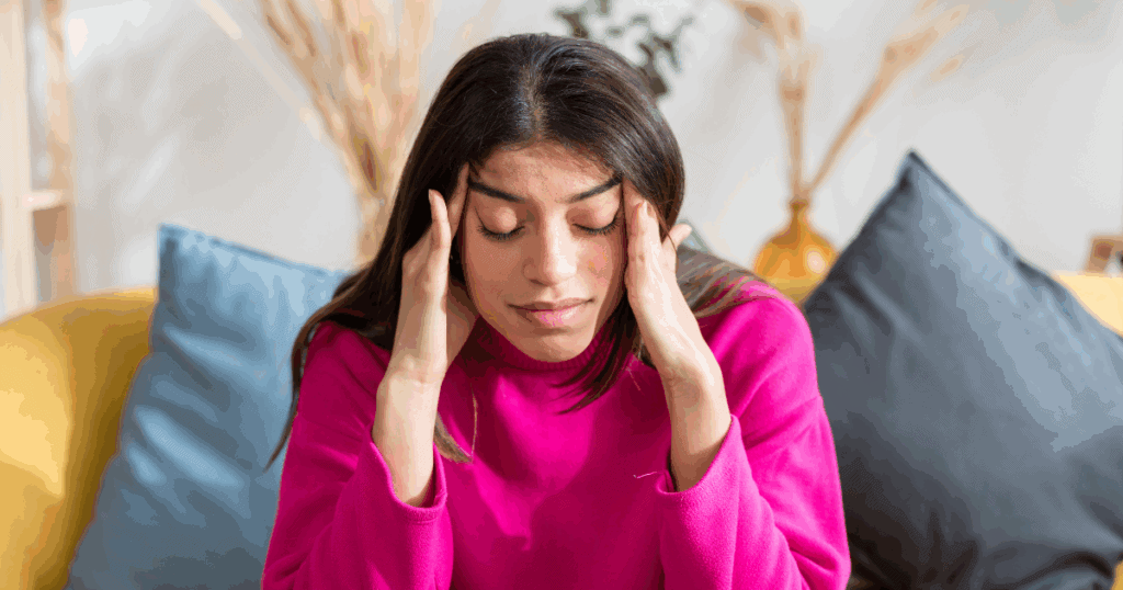 young woman with dark hair in a pink sweater sitting on a couch holding her head due to headaches and migraines